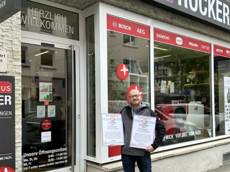 Herr Dengler steht vor dem Rocker Service Mainz Hausgeräte Fachgeschäft mit Eingang und Auszeichnungen, Fachberater für Hausgeräte, Service und Reparaturen in Mainz.