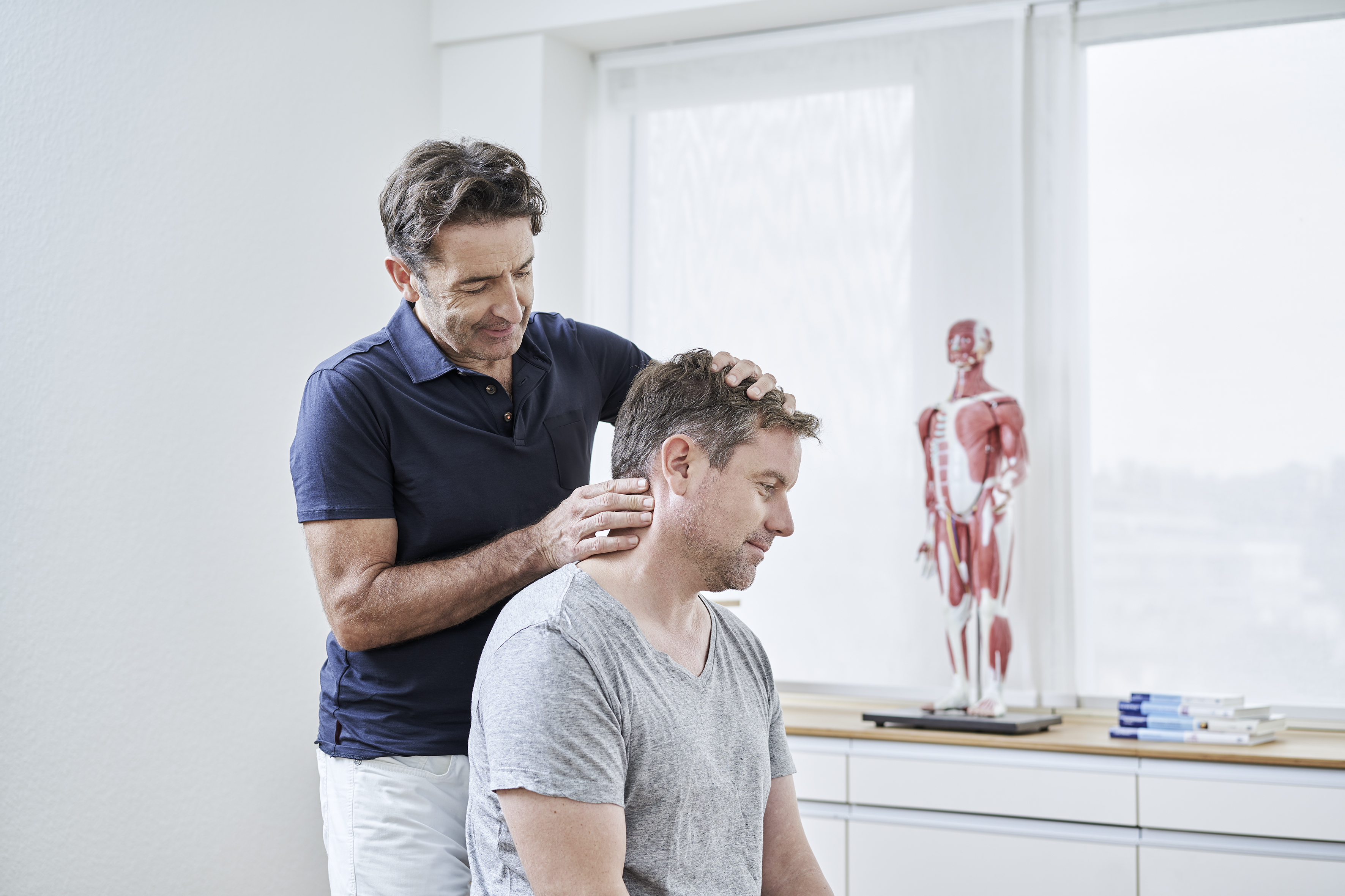 Dr. Gassen behandelt sitzenden Mann manuell am Nacken in medizinischer Praxis mit Anatomie-Modell