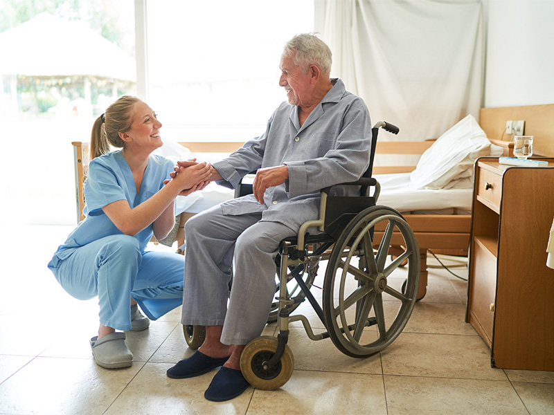 Pflegekraft hält die Hand eines älteren Mannes im Rollstuhl in seinem Zimmer und begleitet ihn fürsorglich bei der Betreuung.